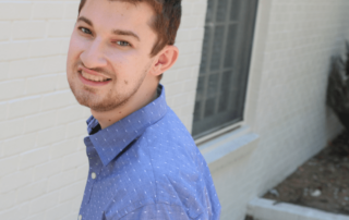 White male student outside, wearing a blue button up collard shirt, in front of a white brick wall.