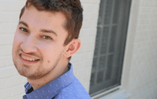 White male student outside, wearing a blue button up collard shirt, in front of a white brick wall.