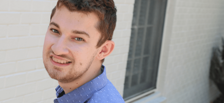 White male student outside, wearing a blue button up collard shirt, in front of a white brick wall.
