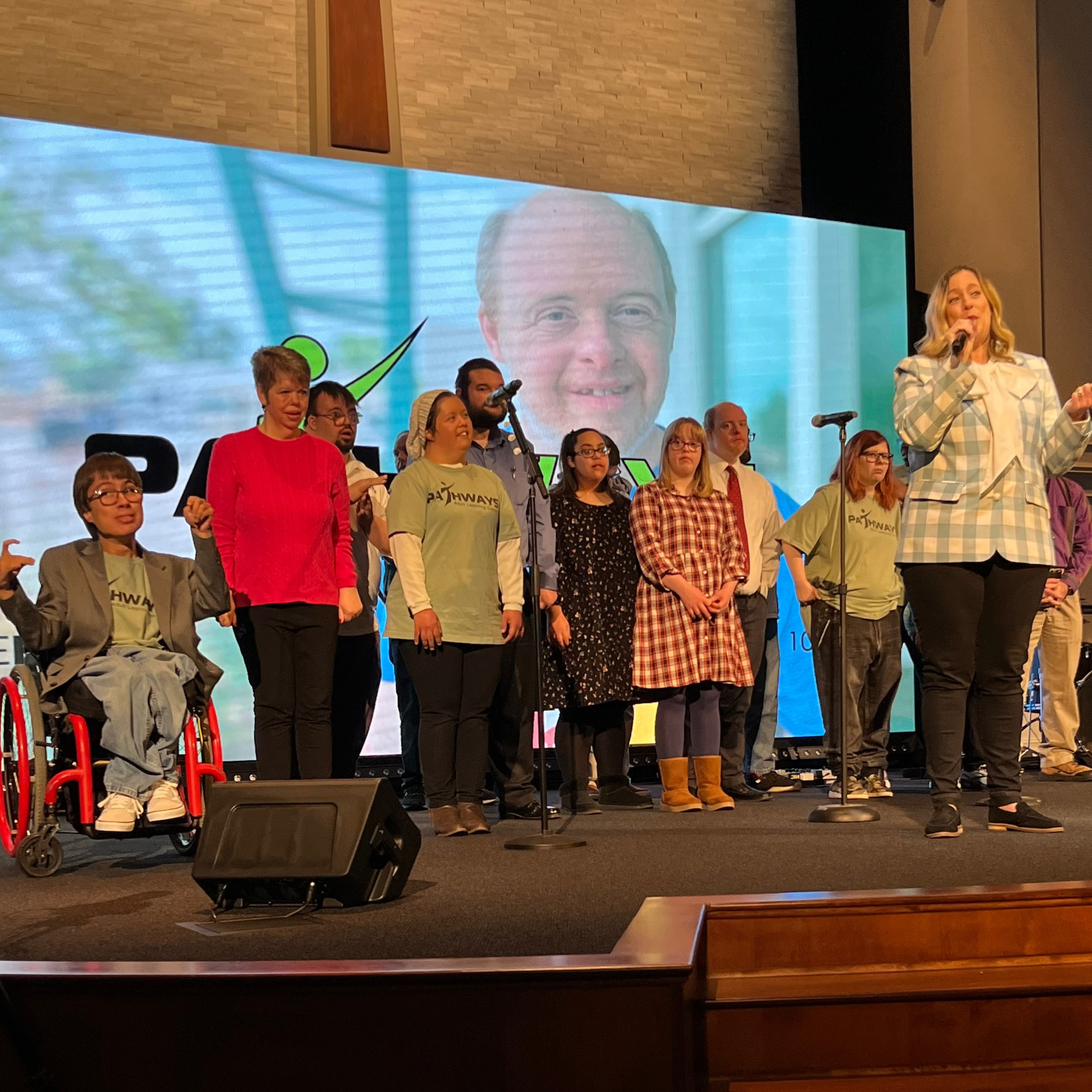 A group of Pathways students on a stage with a Monique, Executive Director, addressing the crowd.