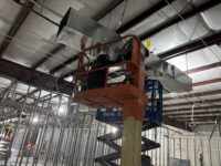 A construction worker on a lift close to the ceiling of building holding piece of metal duct work.