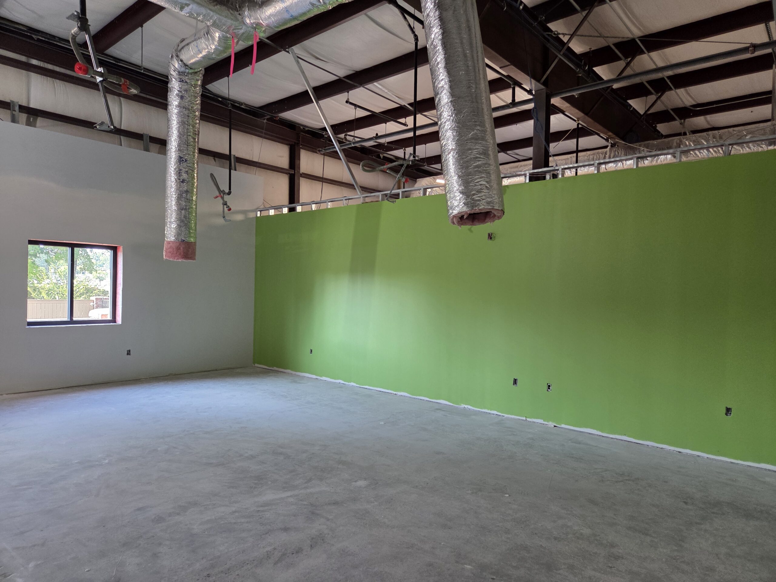 A room with a window and green wall. Air ducts are hanging from the empty ceiling.